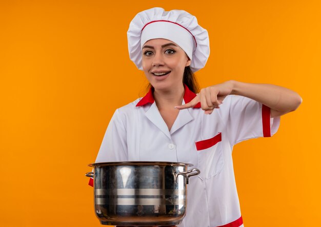 Jovem alegre caucasiana cozinheira com uniforme de chef segura e aponta para a panela isolada na parede laranja com espaço de cópia