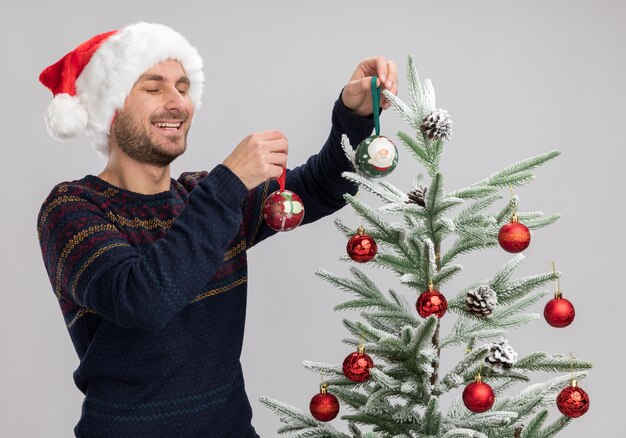 Jovem alegre, caucasiano, usando um chapéu de Natal em pé perto da árvore de Natal, decorando-o com bolas de enfeite de natal, rindo com os olhos fechados, isolado no fundo branco