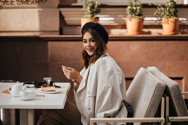 Jovem alegre com cabelo escuro, boina, gabardine bege clássico, sentada à mesa do terraço do café da cidade, sorrindo, tomando cheesecake e chá no café da manhã