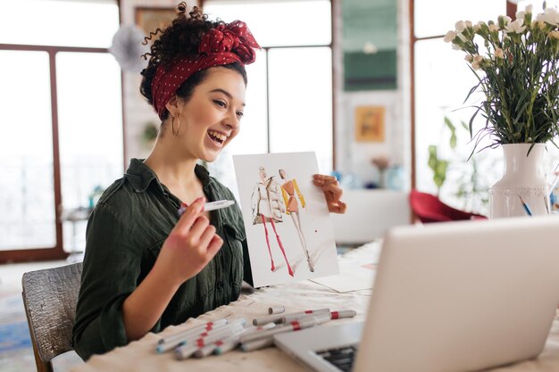 Jovem alegre com cabelos cacheados escuros, sentado à mesa mostrando alegremente o esboço de moda no laptop enquanto passa o tempo na oficina moderna e aconchegante com grandes janelas