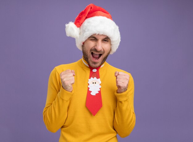 Jovem alegre com chapéu de natal e gravata piscando fazendo gesto de sim isolado na parede roxa com espaço de cópia