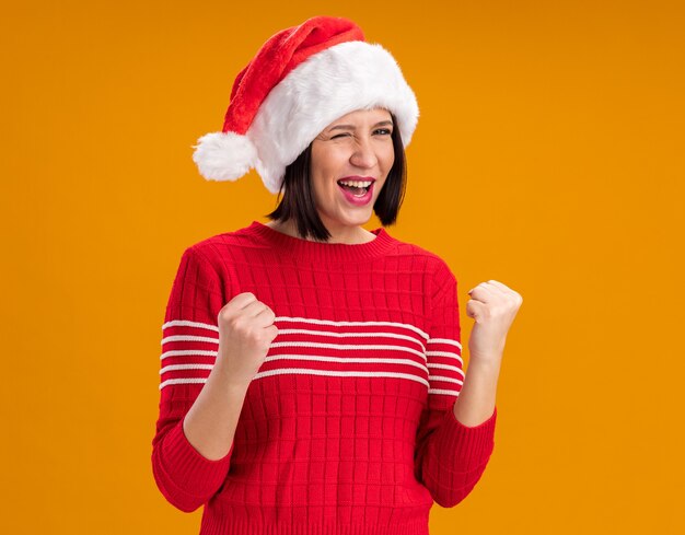 Jovem alegre com chapéu de Papai Noel piscando fazendo gesto de sim isolado na parede laranja