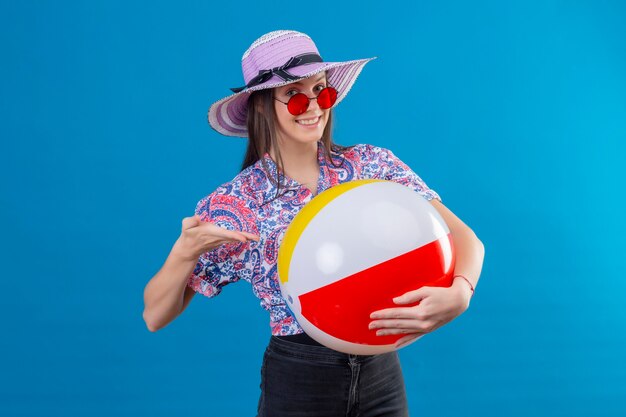 Jovem alegre com chapéu de verão, usando óculos escuros vermelhos, segurando uma bola inflável apontando com o braço da mão para ela, sorrindo com uma cara feliz de pé sobre o espaço azul