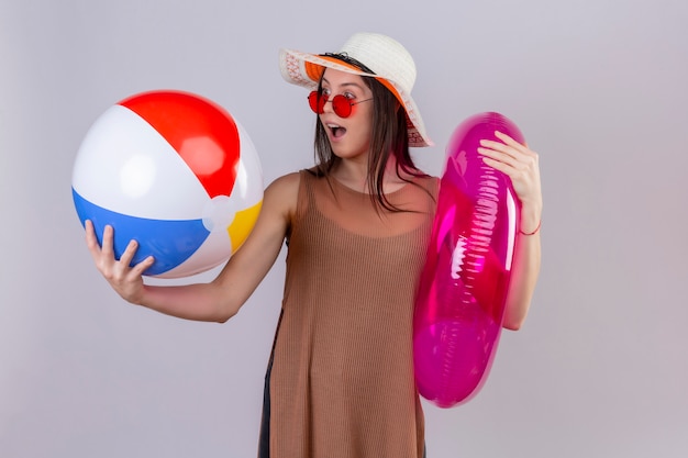 Jovem alegre com chapéu usando óculos escuros vermelhos segurando um anel inflável e uma bola, olhando para ele surpreso e feliz em pé no branco