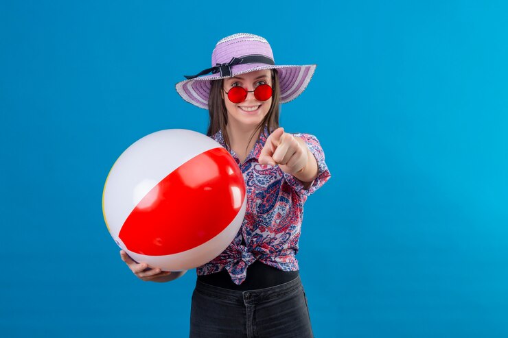 Foto grátis jovem alegre com chapéu usando óculos escuros vermelhos segurando uma bola inflável apontando com o dedo e sorrindo em pé no azul