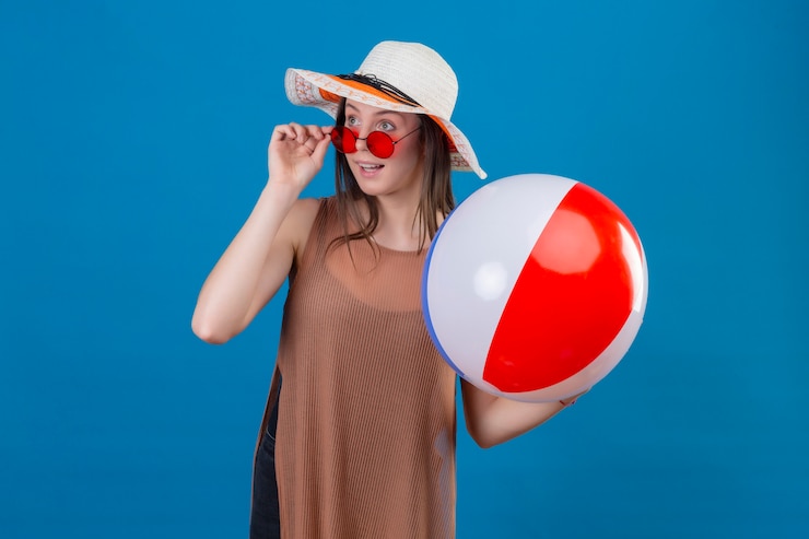 Foto grátis jovem alegre com chapéu usando óculos escuros vermelhos segurando uma bola inflável olhando para o lado com um sorriso feliz parado no azul