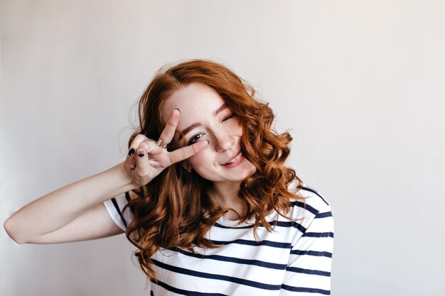 Jovem alegre com pele pálida e cabelo ruivo, posando com o símbolo da paz. Garota incrível em t-shirt listrada isolada.