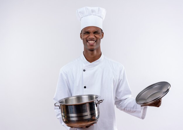 Jovem alegre cozinheira afro-americana com uniforme de chef segurando uma panela na parede branca isolada