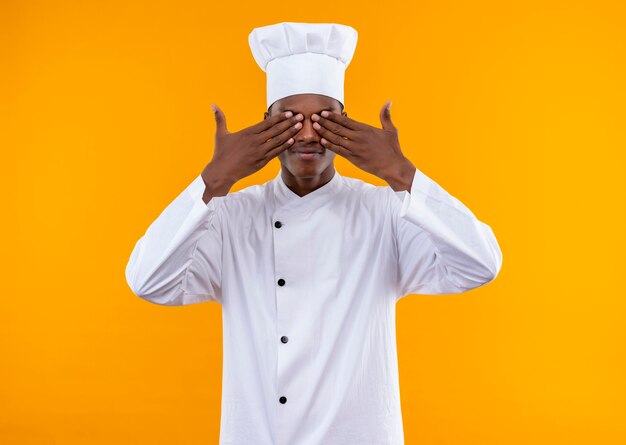 Jovem alegre cozinheira afro-americana em uniforme de chef fecha os olhos com as mãos isoladas na parede laranja