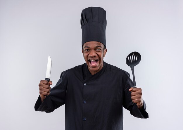 Jovem alegre cozinheira afro-americana em uniforme de chef segura uma faca e uma espátula isoladas na parede branca