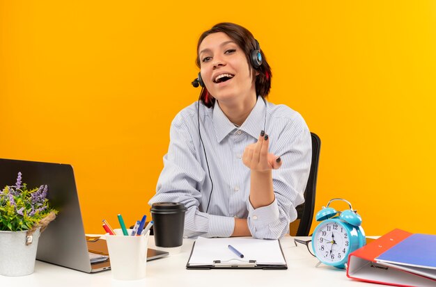 Foto grátis jovem alegre de call center usando fone de ouvido, sentada na mesa, fazendo gesto de venha cá isolado em laranja