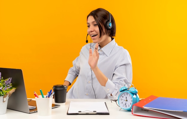 Foto grátis jovem alegre de call center usando fone de ouvido, sentada na mesa, olhando e acenando para um laptop isolado em laranja