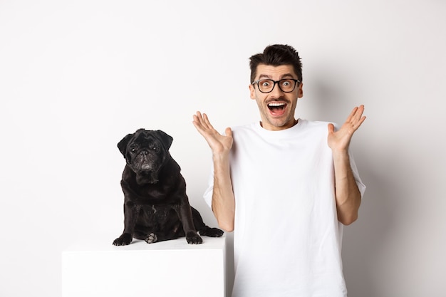 Jovem alegre de óculos em pé com seu animal de estimação, regozijando-se e olhando para a câmera divertido, ouve boas notícias, em pé com um pug sobre fundo branco