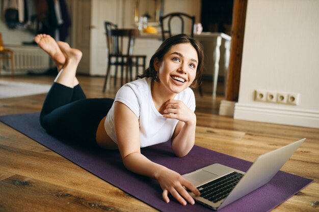 Jovem alegre de olhos azuis e tamanho em roupas esportivas, deitada no tapete de ioga na frente do laptop aberto, relaxando após os exercícios físicos.