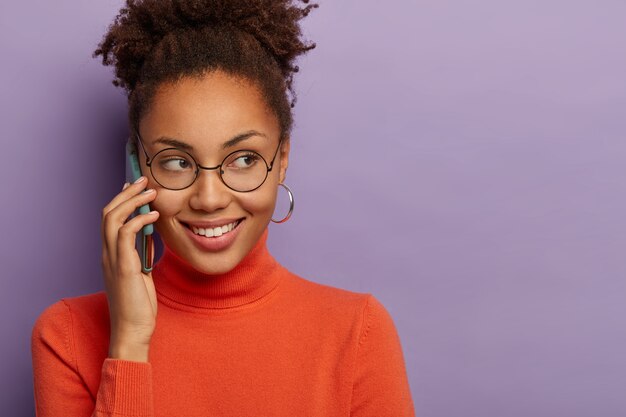 Jovem alegre de pele escura, feminina, conversando no celular, usa óculos redondos transparentes, tem um sorriso encantador, ouve boas notícias, isolada na parede roxa do estúdio, copie a área