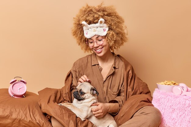 Jovem alegre de pijama macio brinca com cachorro favorito na cama acorda tarde tem fim de semana preguiçoso em casa isolado sobre fundo marrom Modelo feminino de cabelos cacheados feliz expressa amor ao animal de estimação