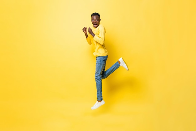 Jovem alegre e animado africano pulando com as mãos apertadas no fundo do estúdio amarelo isolado