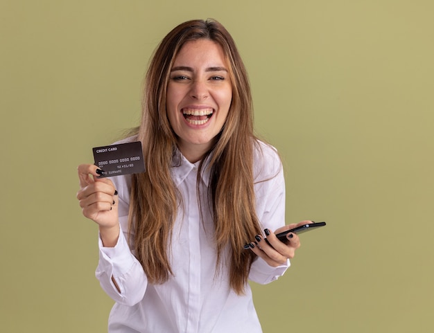 Foto grátis jovem alegre e bonita caucasiana segurando um cartão de crédito e um telefone