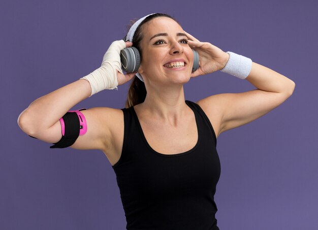 Jovem alegre e bonita e esportiva usando pulseira, fones de ouvido e uma braçadeira de telefone com o pulso ferido enrolado em uma bandagem olhando para cima agarrando os fones de ouvido