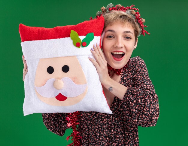 Jovem alegre e bonita usando coroa de flores de Natal e guirlanda de ouropel em volta do pescoço segurando a almofada de Papai Noel e tocando a cabeça com ela isolada na parede verde