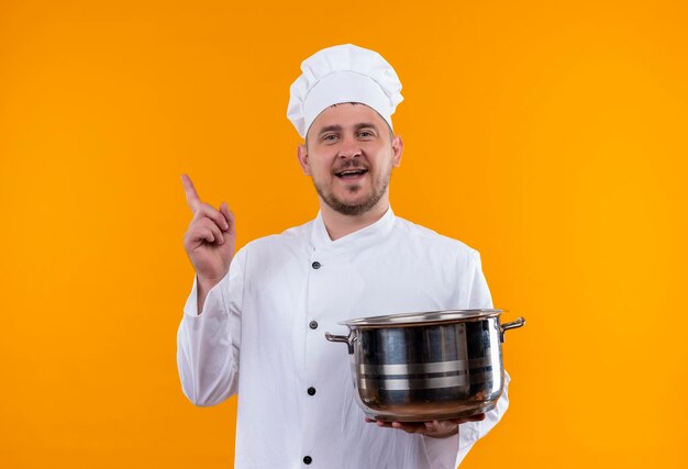 Jovem alegre e bonito cozinheiro em uniforme de chef segurando a panela e levantando o dedo isolado na parede laranja