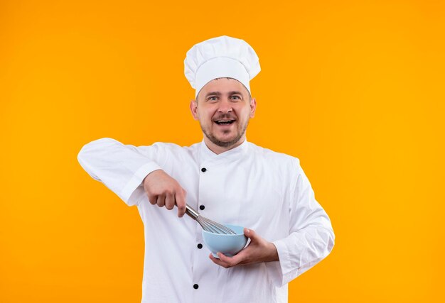 Jovem alegre e bonito cozinheiro em uniforme de chef segurando um batedor e uma tigela isolados na parede laranja