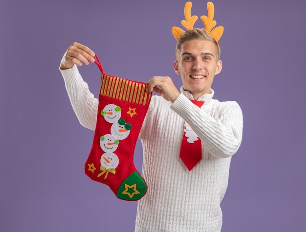 Jovem alegre e bonito usando bandana de chifres de rena e gravata de Papai Noel segurando uma meia de Natal, olhando para a câmera isolada no fundo roxo