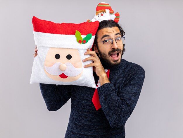 Jovem alegre e bonito usando gravata de Natal com uma argola de cabelo segurando a almofada de Natal ao redor do rosto isolado na parede branca