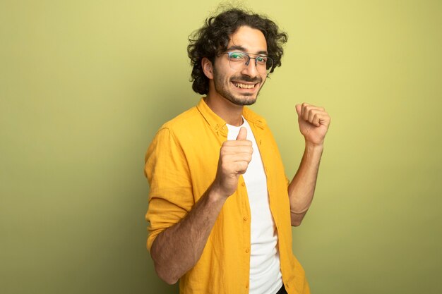 Jovem alegre e bonito usando óculos, em vista de perfil, olhando para a frente, apontando para si mesmo, isolado na parede verde oliva