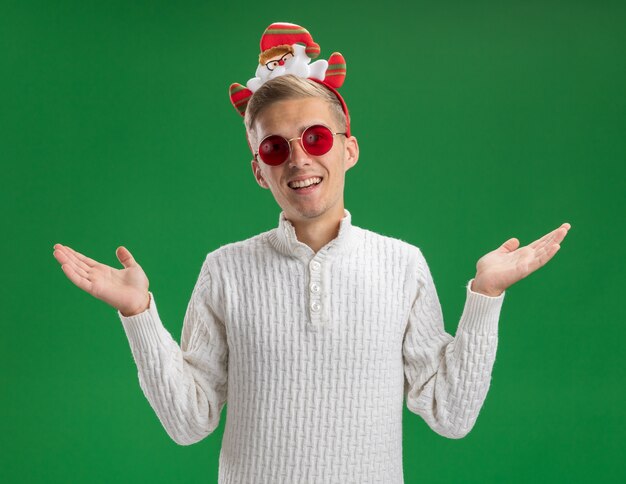 Jovem alegre e bonito usando uma bandana de Papai Noel com óculos, mostrando as mãos vazias, isoladas na parede verde