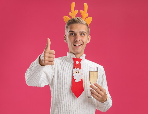 Jovem alegre e bonito usando uma faixa de chifres de rena e gravata de Papai Noel segurando uma taça de champanhe aparecendo o polegar isolado na parede rosa com espaço de cópia