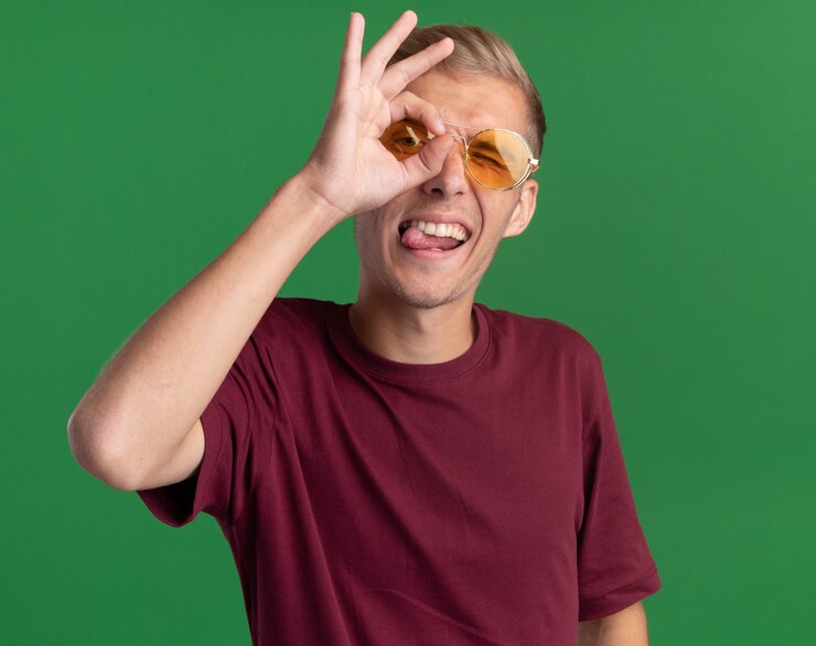 Foto grátis jovem alegre e bonito vestindo camisa vermelha e óculos, mostrando gesto de olhar isolado na parede verde