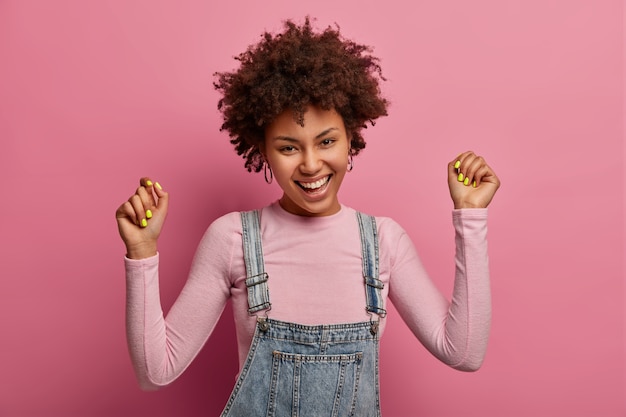 Jovem alegre e cacheada levanta os braços, fica em forma, sorri com alegria, vestida com roupas casuais, estando em alto astral, ri alto, posa contra a parede rosa pastel, se sente a vencedora