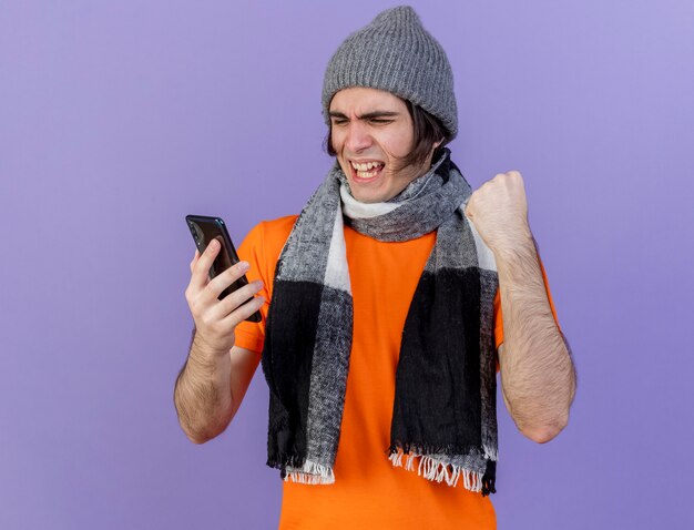 Jovem alegre e doente com chapéu de inverno com lenço segurando e olhando para o telefone, mostrando um gesto de sim isolado no roxo