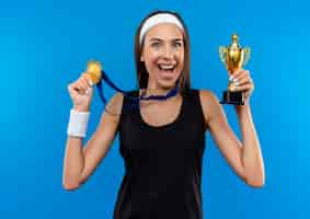 Foto grátis jovem alegre e muito esportiva usando bandana, pulseira e medalha no pescoço, segurando a taça do vencedor e medalha isoladas no espaço azul