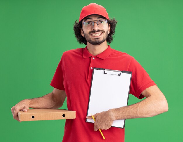 Jovem alegre entregador caucasiano de uniforme vermelho e boné de óculos segurando um lápis de pacote de pizza, mostrando a área de transferência para a câmera