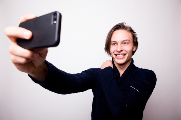 Jovem alegre está tirando uma selfie com um smartphone preto, ele está vestindo uma camisa cinza contra uma parede branca