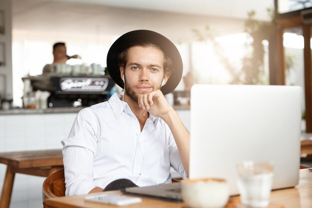 Jovem alegre fazendo uma pausa no café, sentado à mesa com dispositivos eletrônicos e curtindo música online em fones de ouvido