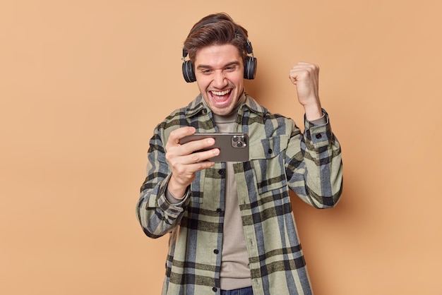 Jovem alegre, feliz por ganhar uma corrida no jogo online, segura o celular horizontalmente, fecha os punhos e se regozija, a vitória exclama de alegria, usa fones de ouvido vestidos com uma camisa xadrez posa dentro de casa