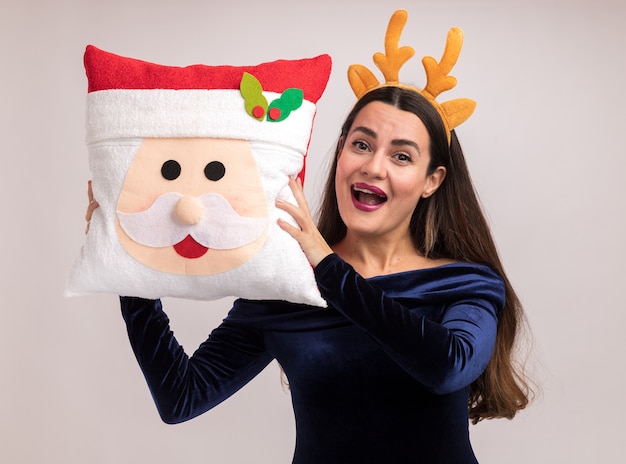 Jovem alegre linda com um vestido azul e uma argola de cabelo de Natal segurando a almofada de Natal ao redor do rosto, isolado na parede branca