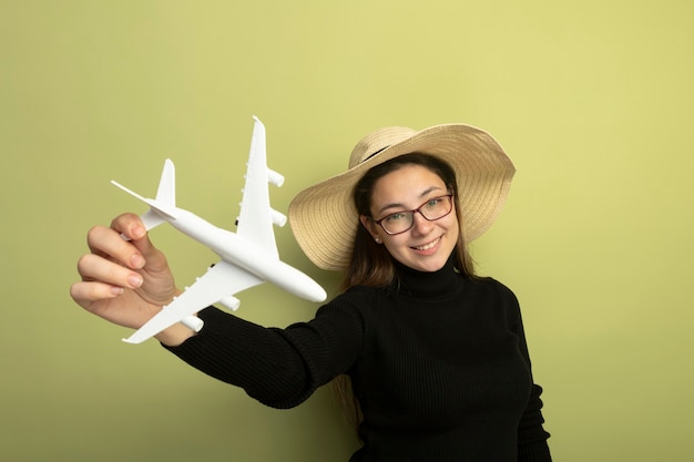 Jovem alegre linda em uma gola preta e óculos segurando um avião de brinquedo sorrindo alegremente feliz e positivo