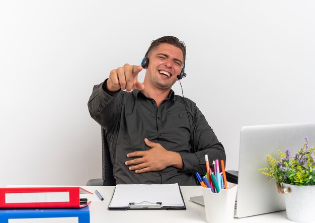 Foto grátis jovem alegre loira trabalhador de escritório com fones de ouvido se senta à mesa com ferramentas de escritório, usando o laptop aponta para a câmera