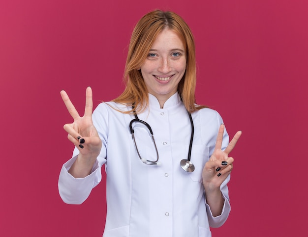 Jovem alegre médica ruiva vestindo túnica médica e estetoscópio fazendo o sinal da paz isolado na parede carmesim