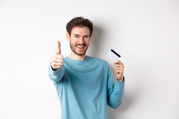 Jovem alegre mostrando o cartão de crédito de plástico e o polegar para cima, gosta e aprova o serviço do bom banco, de pé sobre um fundo branco.