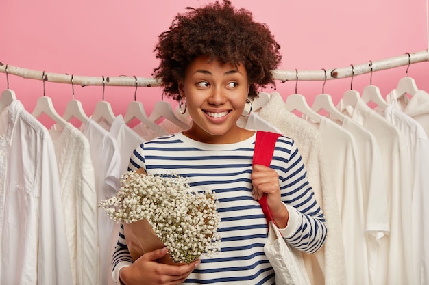 Jovem alegre passa o tempo livre no shopping, olha para o lado com um largo sorriso, carrega uma bolsa de tecido, fica sobre roupas brancas penduradas na fileira, isoladas no fundo rosa.