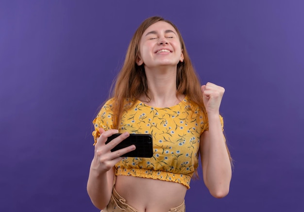 Jovem alegre segurando um telefone celular e levantando o punho com os olhos fechados em um espaço roxo isolado com espaço de cópia