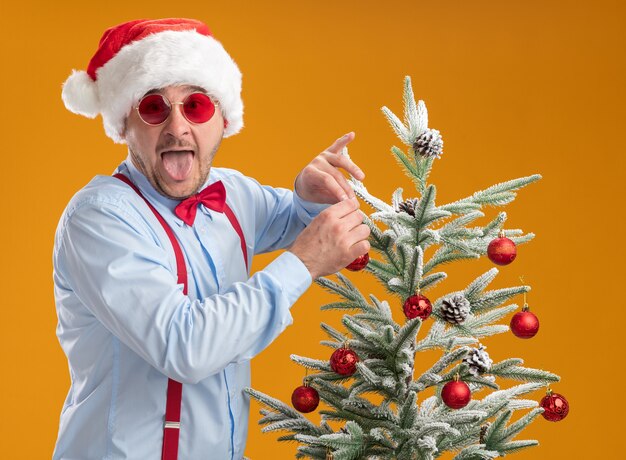 Jovem alegre usando suspensórios gravata borboleta com chapéu de papai noel e óculos vermelhos em pé perto da árvore de natal pendurando o brinquedo na árvore feliz e surpreso mostrando a língua sobre um fundo laranja