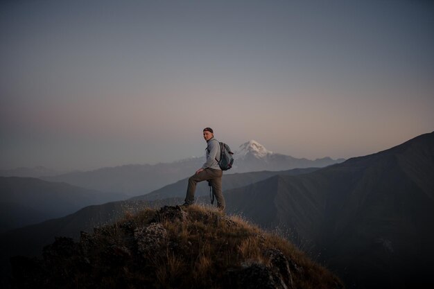 Jovem alpinista caucasiano parado no topo de uma montanha gramada e admirando a natureza nebulosa