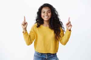 Foto grátis jovem aluna alegre e otimista com penteado encaracolado escuro, suéter amarelo, sorrindo e rindo alegremente, apontando o dedo para cima, mostrando aos amigos o link para o site ou copie o espaço, parede branca