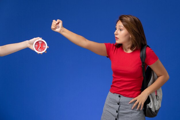 Jovem aluna de camisa vermelha, usando mochila de vista frontal, ameaça sobre fundo azul claro.